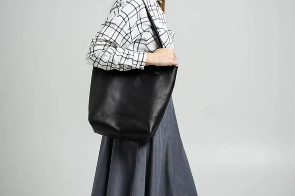 Small black leather bag in a womans hand on a white background. Shoulder handbag. Woman in a white plaid shirt and black jeans and with a black handbag. Style, retro, fashion, vintage and elegance.