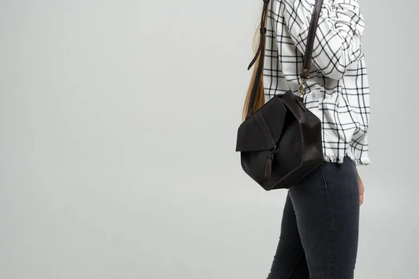 Small black leather bag in a womans hand on a white background. Shoulder handbag. Woman in a white plaid shirt and black jeans and with a black handbag. Style, retro, fashion, vintage and elegance. — Stock Photo, Image