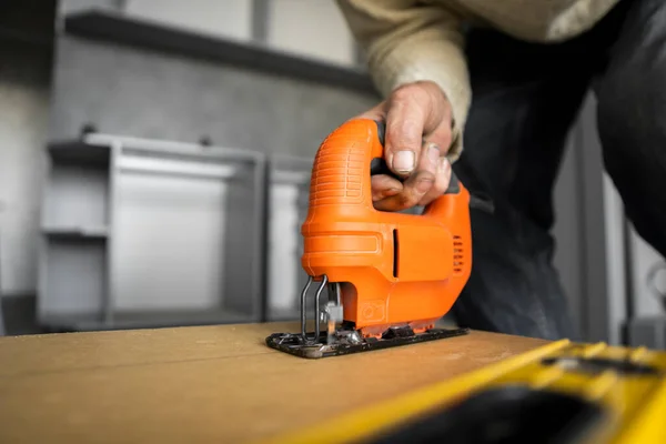 Man hand using electric jigsaw for cutting a fiberboard on a back side of a custom kitchen cabinet. Kitchen installation. Carpenter with a professional hand electric jigsaw instrument.
