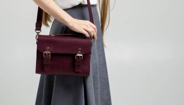 Beyaz arka planda bir kadının omzunun üzerinde küçük kırmızı deri bir çanta. Omuz çantası. Beyaz gömlekli, gri etekli ve kırmızı çantalı bir kadın. Tarz, retro, moda, klasik ve zarafet.