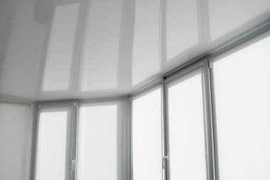 Oturma odasındaki bir balkonun plastik camında beyaz kumaş jaluzi ve esnek bir tavan yansıması var..