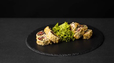 Taze somon, avokado, ananas soslu masago havyarı ve siyah bir masa ve arka plandaki siyah bir tabağa süsleme olarak salata serpiştirilmiş özel suşi rulosu..