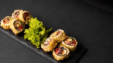 Taze somon, avokado, ananas soslu masago havyarı ve siyah bir masa ve arka plandaki siyah bir tabağa süsleme olarak salata serpiştirilmiş özel suşi rulosu..