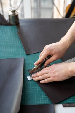 Erkekler ellerinde kırtasiye bıçağı ve metal cetvelle atölyesinde deri bir cüzdan için parça kesiyorlar. Kahverengi doğal deri ile çalışma süreci. Bir zanaatkar elinde zanaat araçları tutuyor..
