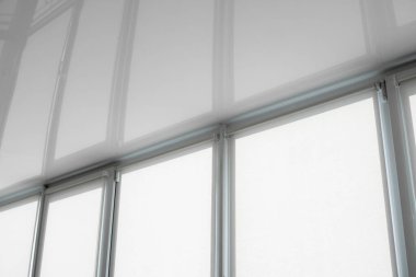 Oturma odasındaki bir balkonun plastik camında beyaz kumaş jaluzi ve esnek bir tavan yansıması var..