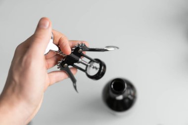 Arka planda açık beyaz bir mutfağı olan kırmızı şarap şişesi olan bir adamın elindeki metal tirbuşon. Şarap aşığı. Şarap açacağı..