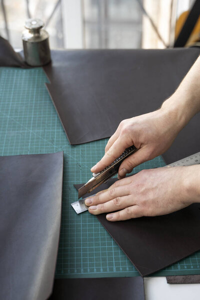 Mens hand holding a stationery knife and metal ruler and cutting on a pieces for a leather wallet in his workshop. Working process with a brown natural leather. Craftsman holding a crafting tools.