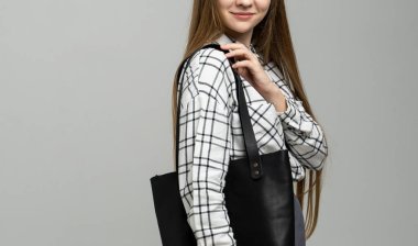 Beyaz arka planda bir kadının elindeki küçük siyah deri çanta. Omuz çantası. Beyaz ekose gömlekli, siyah kotlu ve siyah çantalı bir kadın. Tarz, retro, moda, klasik ve zarafet.