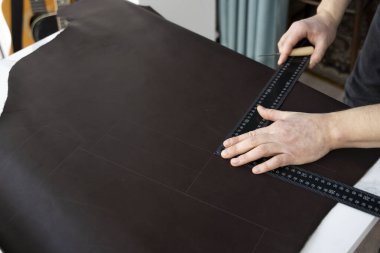 Erkekler bir teneke kutu ve metal cetvel tutuyor ve atölyesinde deri bir cüzdan üzerinde çalışıyorlar. Kahverengi doğal deri ile çalışma süreci. Zanaatkar elinde bir el işi aleti tutuyor ve deriye işaretler koyuyor..