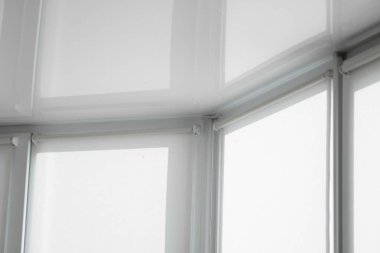 Oturma odasındaki bir balkonun plastik camında beyaz kumaş jaluzi ve esnek bir tavan yansıması var..