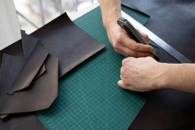 Kırtasiye bıçağı ve metal cetvel tutan ve atölyesinde deri bir cüzdan için parça kesen adamlar. Kahverengi doğal deri ile çalışma süreci. Bir zanaatkar elinde zanaat araçları tutuyor..