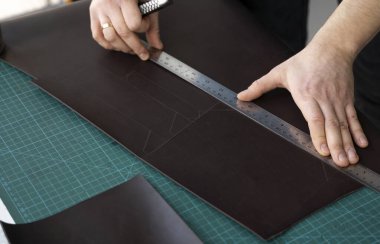 Kırtasiye bıçağı ve metal cetvel tutan ve atölyesinde deri bir cüzdan için parça kesen adamlar. Kahverengi doğal deri ile çalışma süreci. Bir zanaatkar elinde zanaat araçları tutuyor..
