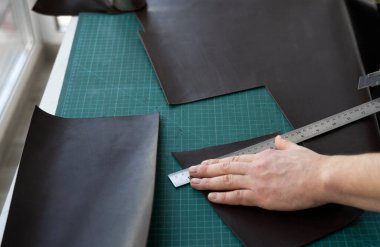 Kırtasiye bıçağı ve metal cetvel tutan ve atölyesinde deri bir cüzdan için parça kesen adamlar. Kahverengi doğal deri ile çalışma süreci. Bir zanaatkar elinde zanaat araçları tutuyor..