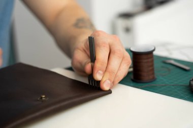 Erkekler deri bir çekiç ve çelik keski ile delik yumruğu tutuyor ve atölyesinde deri bir cüzdan yapıyor. Kahverengi doğal deri ile çalışma süreci. Bir zanaatkar elinde zanaat araçları tutuyor..