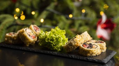 Nori, taze somon, ton balığı, avokado, masago havyarı, ananas sosuyla süslenmiş salata, Noel için ahşap bir masada siyah bir tabağa süsleme olarak serpilmiş özel suşi rulosu..