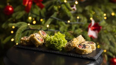 Nori, taze somon, ton balığı, avokado, masago havyarı, ananas sosuyla süslenmiş salata, Noel için ahşap bir masada siyah bir tabağa süsleme olarak serpilmiş özel suşi rulosu..