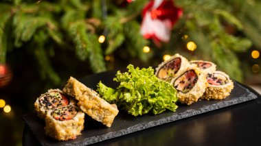 Nori, taze somon, ton balığı, avokado, masago havyarı, ananas sosuyla süslenmiş salata, Noel için ahşap bir masada siyah bir tabağa süsleme olarak serpilmiş özel suşi rulosu..