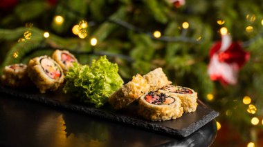 Nori, taze somon, ton balığı, avokado, masago havyarı, ananas sosuyla süslenmiş salata, Noel için ahşap bir masada siyah bir tabağa süsleme olarak serpilmiş özel suşi rulosu..