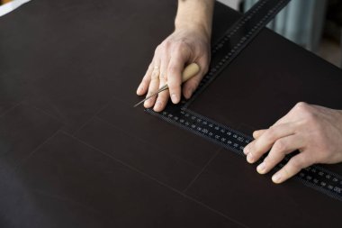 Erkekler bir teneke kutu ve metal cetvel tutuyor ve atölyesinde deri bir cüzdan üzerinde çalışıyorlar. Kahverengi doğal deri ile çalışma süreci. Zanaatkar elinde bir el işi aleti tutuyor ve deriye işaretler koyuyor..