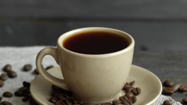 Ein Tropfen Kaffee lässt eine beige Tasse Espresso fallen, Kaffeebohnen liegen auf dem Tisch verstreut und ein Leinentuch auf einem Holztisch. Frischer köstlicher Kaffee steht zum Trinken bereit. — Stockvideo