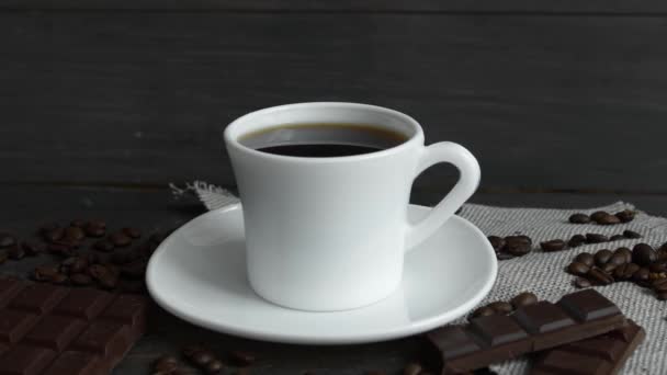 Una gota de café cae en una taza blanca de espresso con granos de café esparcidos en la mesa y chocolate en una mesa de madera. Café fresco delicioso está listo para beber. — Vídeos de Stock