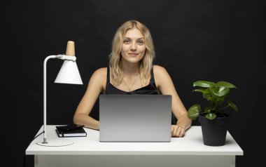 Profesyonel bir kadın, dizüstü bilgisayarı ve lambası ile beyaz masada oturuyor, karanlık arka plana karşı, özgüvenle çalışıyor ve görevine odaklanıyor..