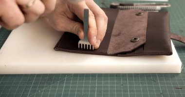 Kahverengi deri cüzdanda delikler açmak için metal delme aleti kullanan zanaatkârın yakın çekimi, geleneksel el yapımı deri işçiliği ve hassasiyeti gösteriyor.