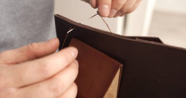 Craftsman hands sewing brown leather wallet using needle and thread. Emphasizes handmade quality, durability, and traditional leatherworking skills