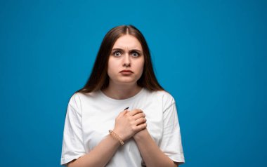 Mavi gözlü, uzun kahverengi saçlı, el ele tutuşan korkmuş bir kadının portresi. Beyaz tişört, mavi duvar..