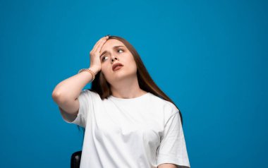 Mavi gözlü, kahverengi saçlı, alnına dokunan bitkin bir kadın. Beyaz tişört, mavi arka plan, hüsran duygusu..