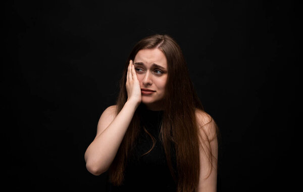 Studio portrait of emotional woman with long brown hair and blue eyes touching face sadly. Wearing black top, dark backdrop.
