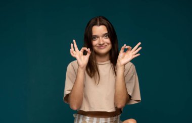 Bej gömlekli neşeli esmer kadın portresi gülümsüyor ve mavi arka planda çift el hareketi gösteriyor.