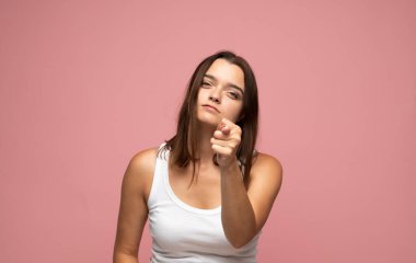Beyaz atletli, kendine güvenen genç esmer kadın kamerayı işaret ederek iddialı duygular gösteriyor ve pembe arka plana odaklanıyor.