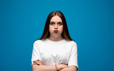 Sinirli genç esmer kadın, mavi arka planda beyaz tişörtlü, tedirgin bir ifadeyle yanakları şişiriyor.