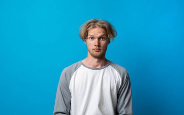 Gri ve beyaz gömlekli genç bir adamın stüdyo portresi. Parlak mavi arka plana karşı geniş gözlü bir şaşkınlık ifadesi..