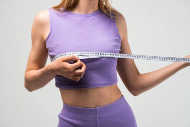 Leylak bluzlu bir kadının stüdyo görüntüsü ve ölçü bandıyla göğsü ölçen şortlar, sağlık, vücut bakımı ve uygunluğu sembolize ediyor..