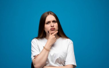 Genç bir kadın şaşkınlık ve şokla tepki verir, temiz mavi stüdyo ortamında kaşlarını kaldırır ve gözlerini genişletir, güçlü duyguların altını çizer..