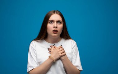 Genç bir kadın geniş gözleri ve elleri göğsünde dramatik bir şekilde tepki veriyor. Parlak mavi bir stüdyo resmine duygusal açıklıkla güçlü bir şok ve güvensizlik gösteriyor..