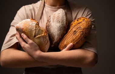 Bir fırıncı üç farklı zanaatkar ekmek somunu tutar, her biri eşsiz dokular ve kabukludur, çeşitlilik ve el yapımı pişirme geleneğini gösterir..