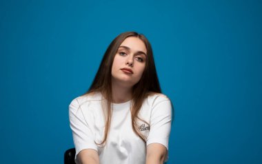 Sakin ve tarafsız görünümlü genç bir kadının rahat bir stüdyo portresi. Yumuşak ışıklandırma ve mavi arka plan temiz, minimalist bir atmosfer yaratır..