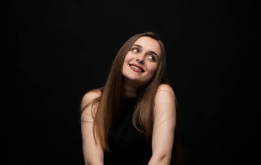 Genç bir kadın, hafif bir stüdyoda, siyah bir arkaplan karşısında ışık tutarak, neşe ve doğal duyguları ifade ederek, yan tarafa bakarken ışıl ışıl gülümsüyor.