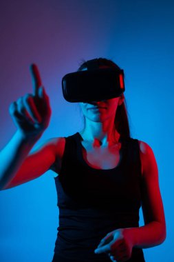 Oyuncu kadın sanal elementlerle etkileşime girerken neon ışıkta bir VR kulaklık takıyor, etkileyici jestlerle dijital alanı keşfediyor.