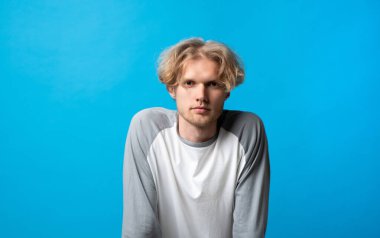 Yakın stüdyo portresi, parlak mavi bir arkaplan karşısında duran ciddi ve odaklanmış bir ifadeyle öne doğru eğilen genç bir adam..