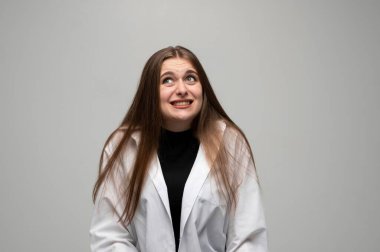 Beyaz önlüklü genç bir kadının portresi gri arka planda garip, komik bir gülümseme yaratıyor. Rahatsızlık, mizah, duygu kavramı.