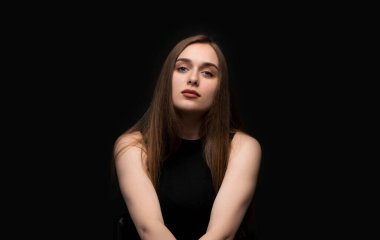 Karanlık bir arka planda nötr bir ifadeyle poz veren genç bir kadının minimalist stüdyo portresi, doğal güzellik ve ruh halini vurguluyor..