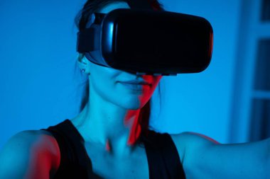 Bir kadın, mavi ve kırmızı ortam aydınlatmasının fütüristik bir karışımı ile çevrili bir VR kulaklıkla sanal bir arayüz keşfediyor..