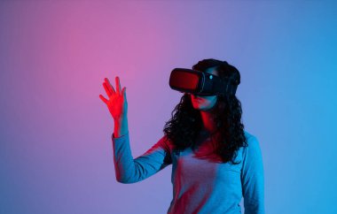 VR kulaklık takan kadın kullanıcı, kasvetli mavi ve kırmızı neon stüdyo ışıklandırmasıyla çevrili dijital sanal arayüzle etkileşime geçmek için uzanıyor..