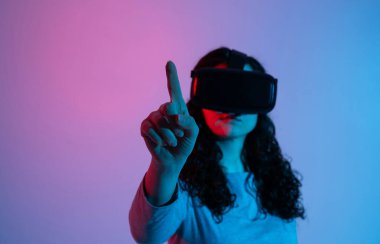 VR kulaklık takan bir kadın, neon mavi ve pembe ışık altında sanal bir arayüzle etkileşime geçerek öne uzanıyor.