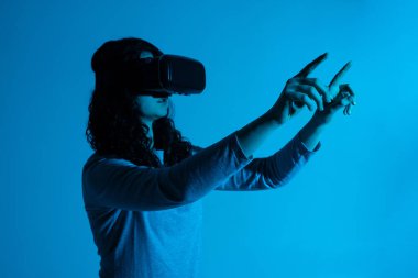 Bir kadın sanal arayüzle etkileşime girer, iki eliyle dışarıya uzanır ve mavi neon ışıklı bir VR kulaklık takar..