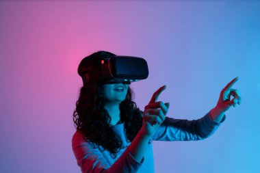 VR kulaklık takan gülümseyen kadın, iki eliyle parlak pembe ve mavi neon ışıklar altında hareket ederek sanal çevreyle etkileşime geçiyor..